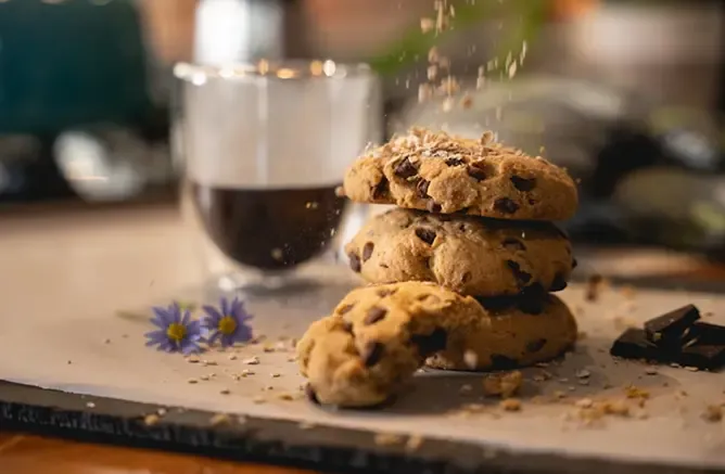 Chocolate chip cookies with sprinkles falling, next to a glass of coffee and flower.