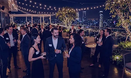 Business professionals networking on a lit rooftop terrace overlooking a vibrant city skyline at night.