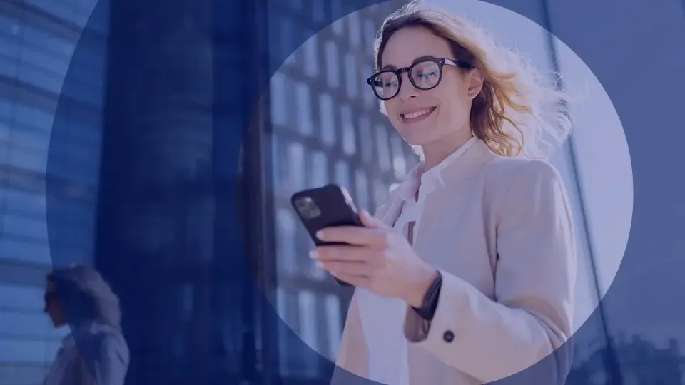 Smiling woman in glasses holds a smartphone outdoors with urban buildings in the background.