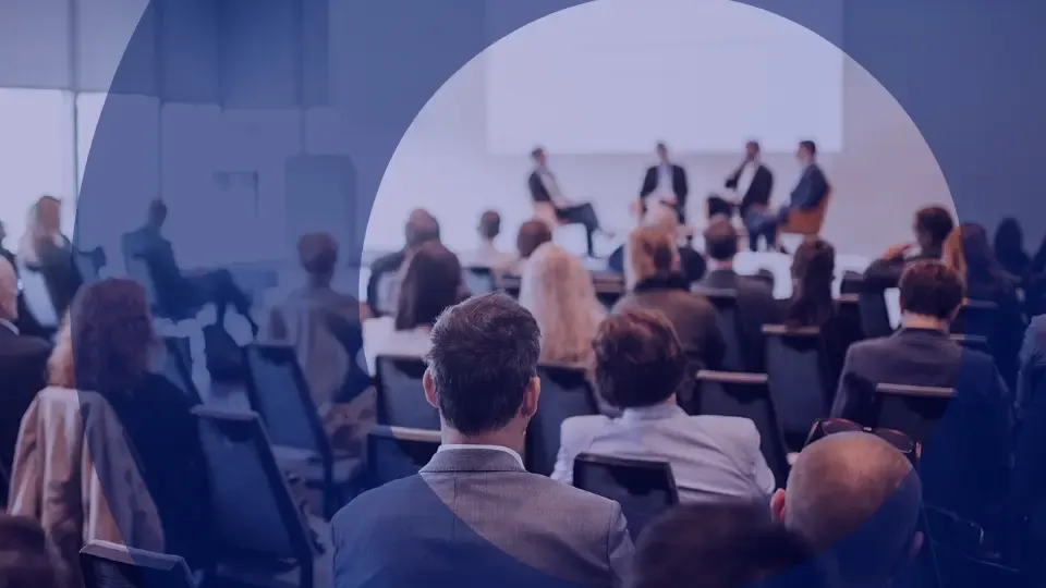 Audience at a conference listens to a panel discussion with speakers on a stage.