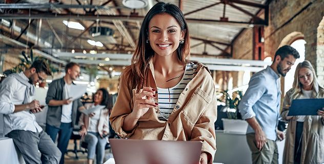 Smiling woman with a laptop in a busy, modern open-plan office.