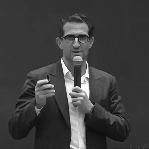 A man in a suit and glasses holds a microphone and gestures while speaking.
