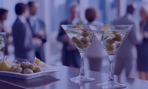 Two martinis with olives and rosemary on a bar, people conversing in background.