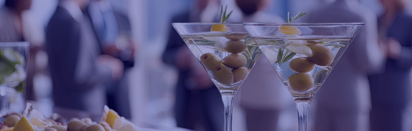 Two martinis with olives and rosemary on a bar, people conversing in background.