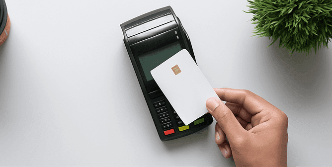Hand tapping a white payment card on a black point-of-sale terminal for contactless payment.