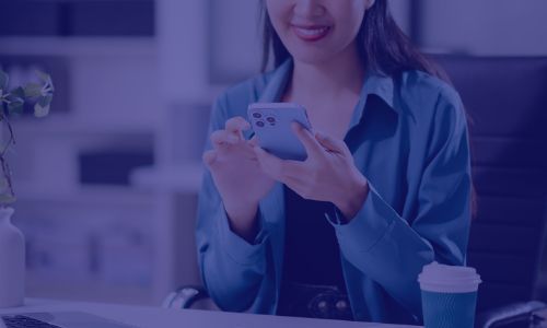 A woman in a blue shirt smiles while looking at her smartphone in an office setting.