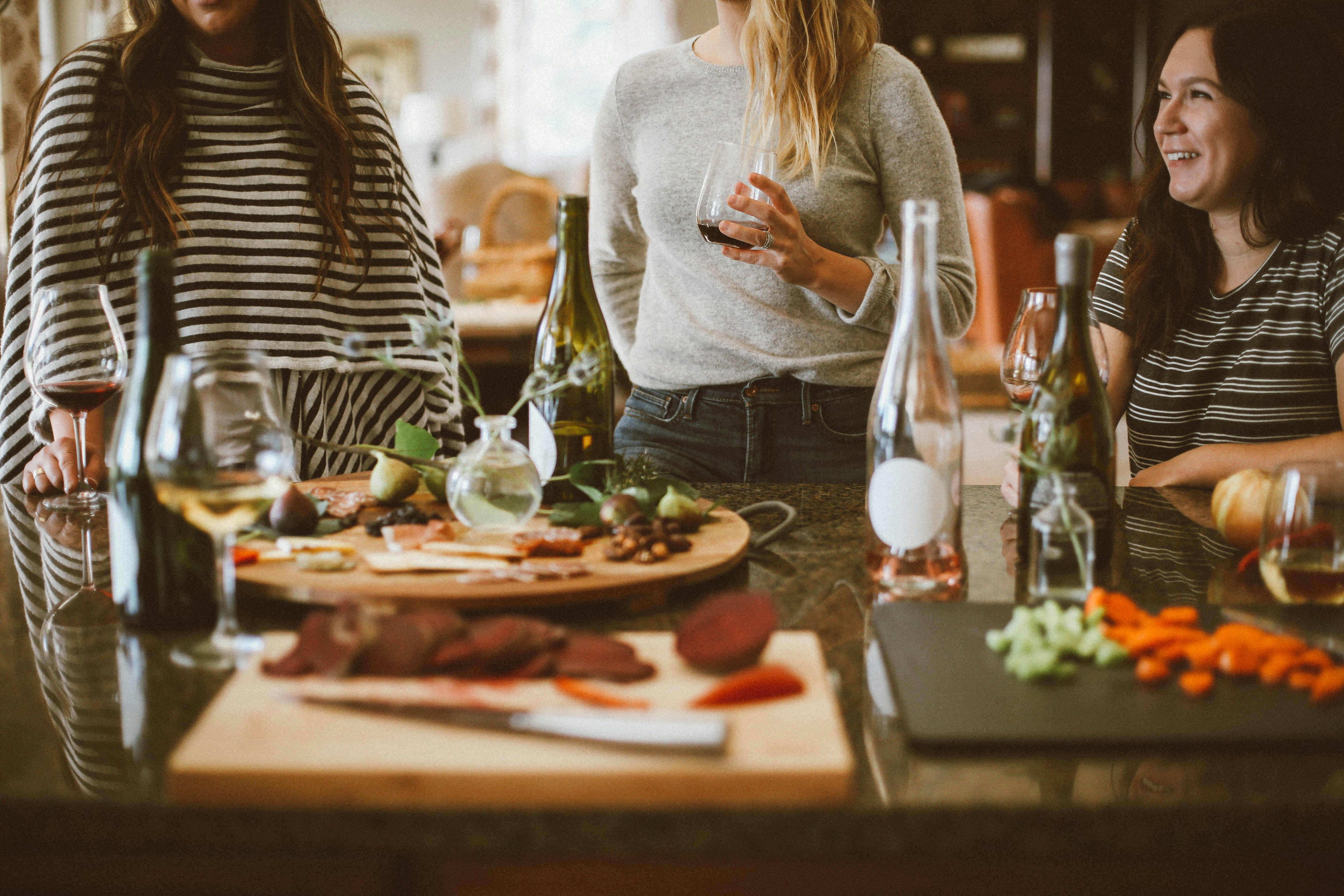 Three women socializing around a kitchen counter with wine, glasses, and food preparations.