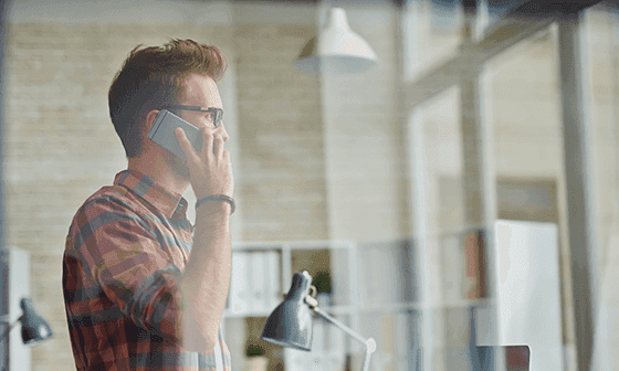 Young man in plaid shirt and glasses talks on phone in a bright office.