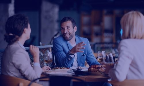 A man in a blue suit jacket gestures while speaking to two women at a restaurant table.
