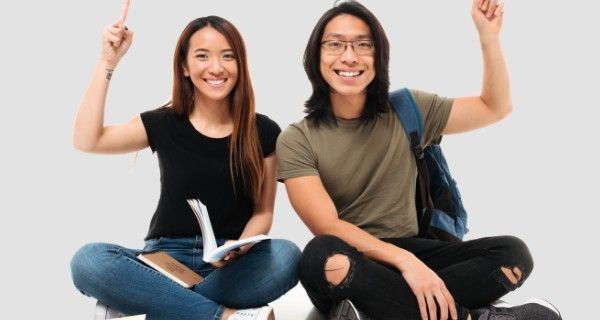 Two smiling Asian students sit on the floor, raising hands with books.
