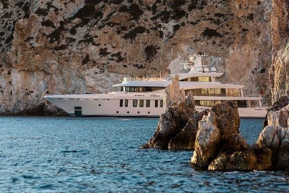Large white yacht anchored in clear blue water next to a rugged rocky cliff.