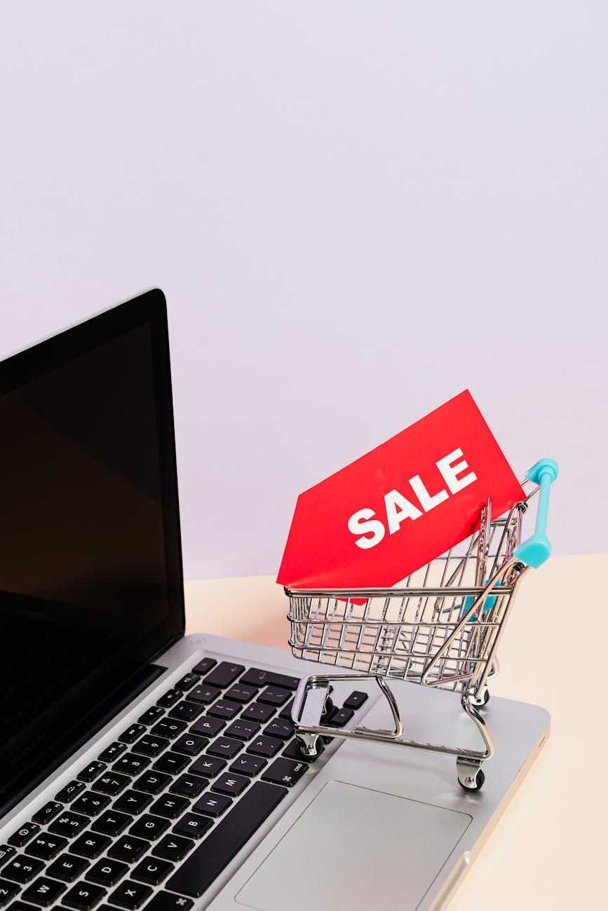 Open laptop next to a miniature shopping cart with a red 'SALE' tag inside.