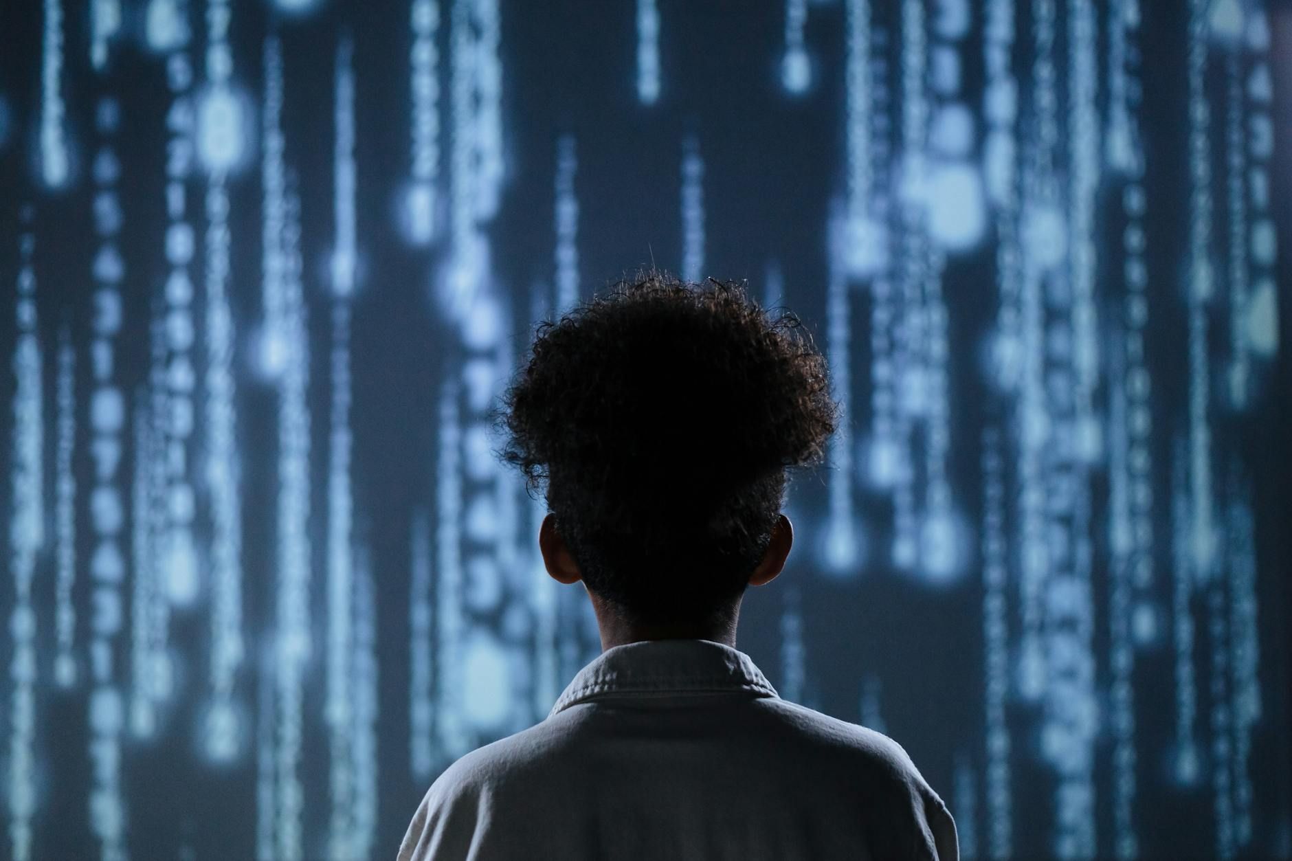 Person from behind observing glowing blue digital lines cascading downwards on a dark screen.