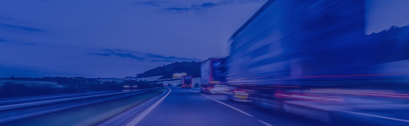 Blurred trucks on a highway at dusk, showing motion and light trails.