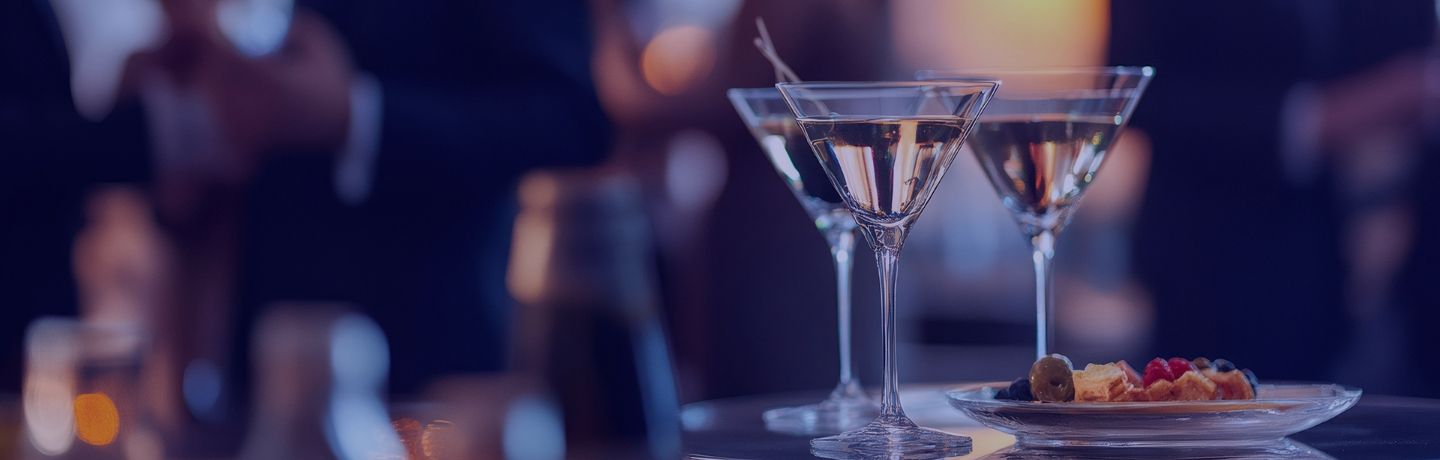 Two martini glasses with drinks and a small snack plate on a reflective bar in a dimly lit setting.